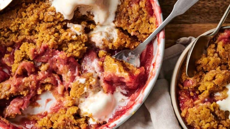 A baked dessert with a crumbly brown topping and red filling sits in a dish, with a portion served in a separate bowl. Spoons are present in both the dish and the bowl.