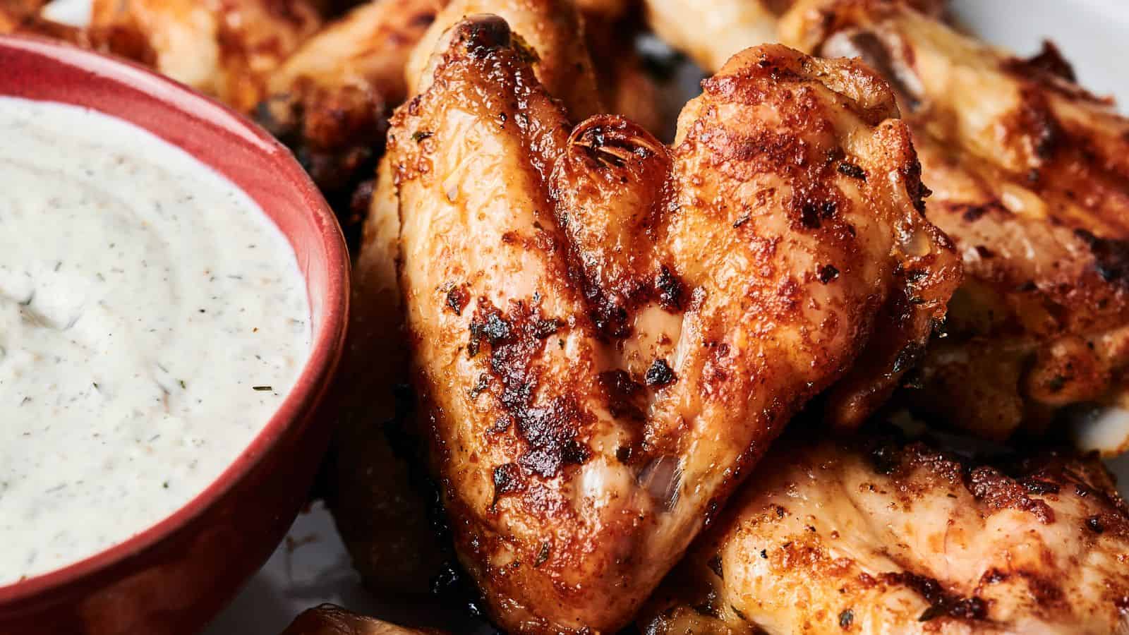 Close-up of grilled chicken wings with a crispy texture, next to a red bowl of creamy dipping sauce.