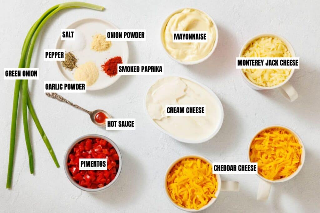 Ingredients for a cheese dip are arranged on a white surface. They include green onion, spices, pimentos, cheese, hot sauce, mayonnaise, and cream cheese.