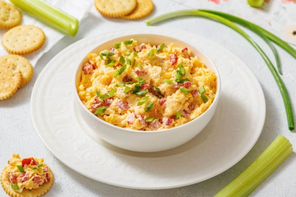 A bowl of creamy pimento cheese topped with chopped green onions sits on a white plate. Crackers and celery sticks surround it on a light surface.