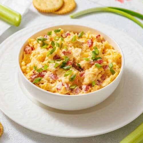 A bowl of creamy pimento cheese topped with chopped green onions sits on a white plate. Crackers and celery sticks surround it on a light surface.
