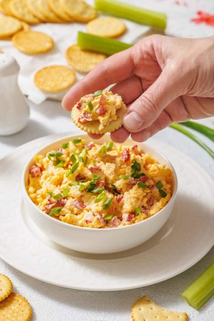 A hand holds a cracker topped with pimento cheese. The bowl of cheese is garnished with green onions. Crackers and celery are in the background.