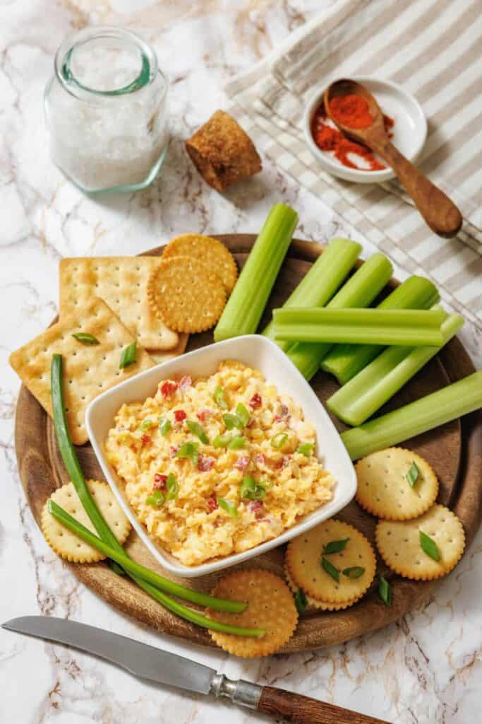 A wooden platter with a bowl of creamy pimento cheese, surrounded by crackers, celery sticks, and green onions. A jar of salt and paprika are nearby.