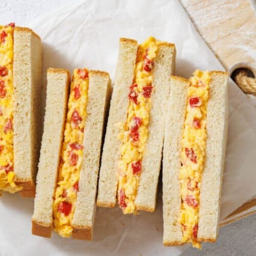 Sandwiches sliced in half on a wooden board, filled with a creamy mix of pimento cheese and red peppers. Presented on parchment paper, evoking a homemade feel.
