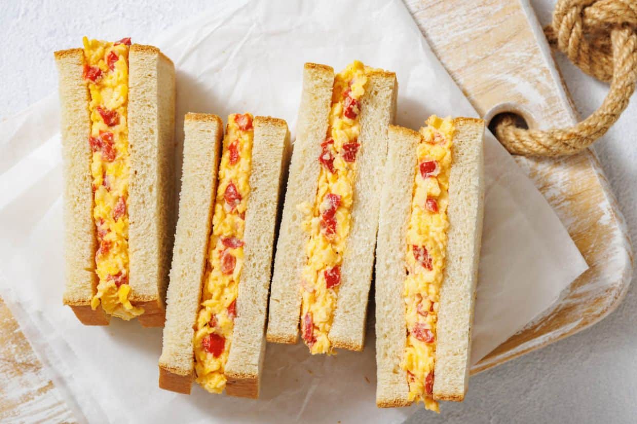 Sandwiches sliced in half on a wooden board, filled with a creamy mix of pimento cheese and red peppers. Presented on parchment paper, evoking a homemade feel.