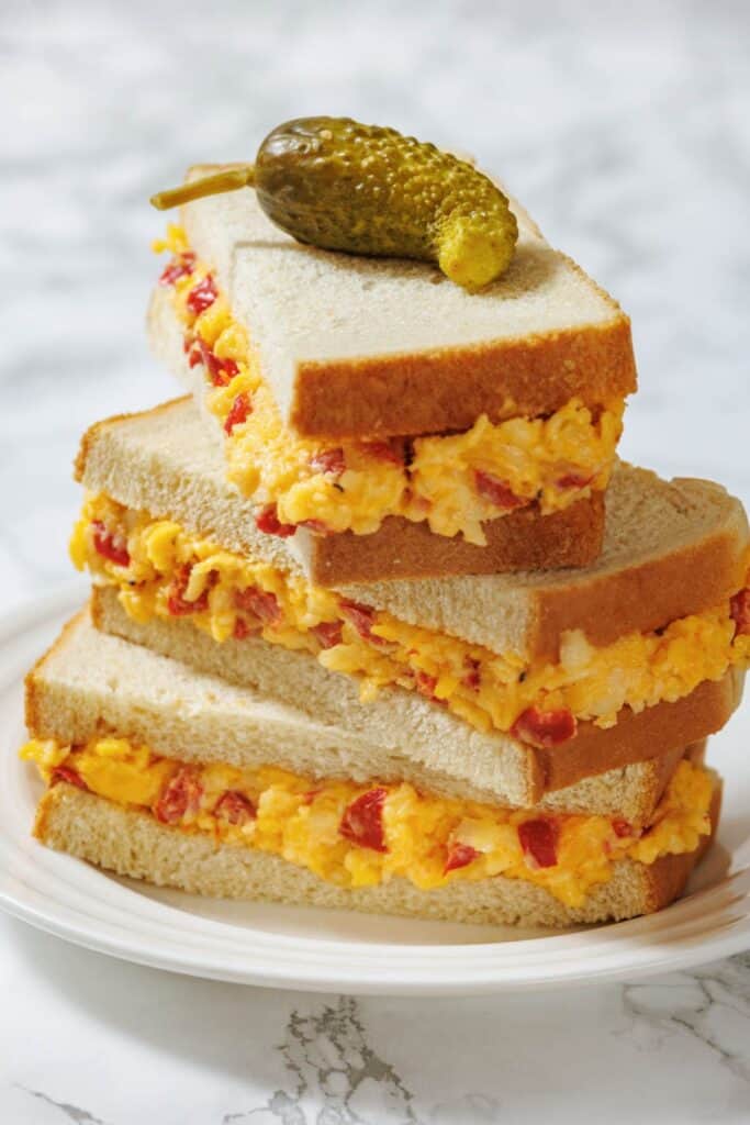 A stack of three pimento cheese sandwiches on white bread, topped with a small pickle, placed on a white plate with a marble background.