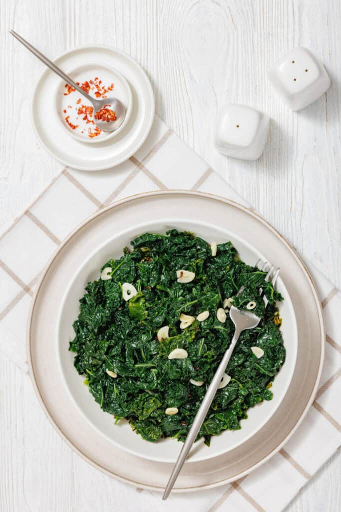 A white bowl filled with sautéed kale, glistening from light oil and garnished with thin slices of garlic. The leafy greens appear vibrant and tender, with a slightly wilted texture, served on a white plate over a light plaid tablecloth background.