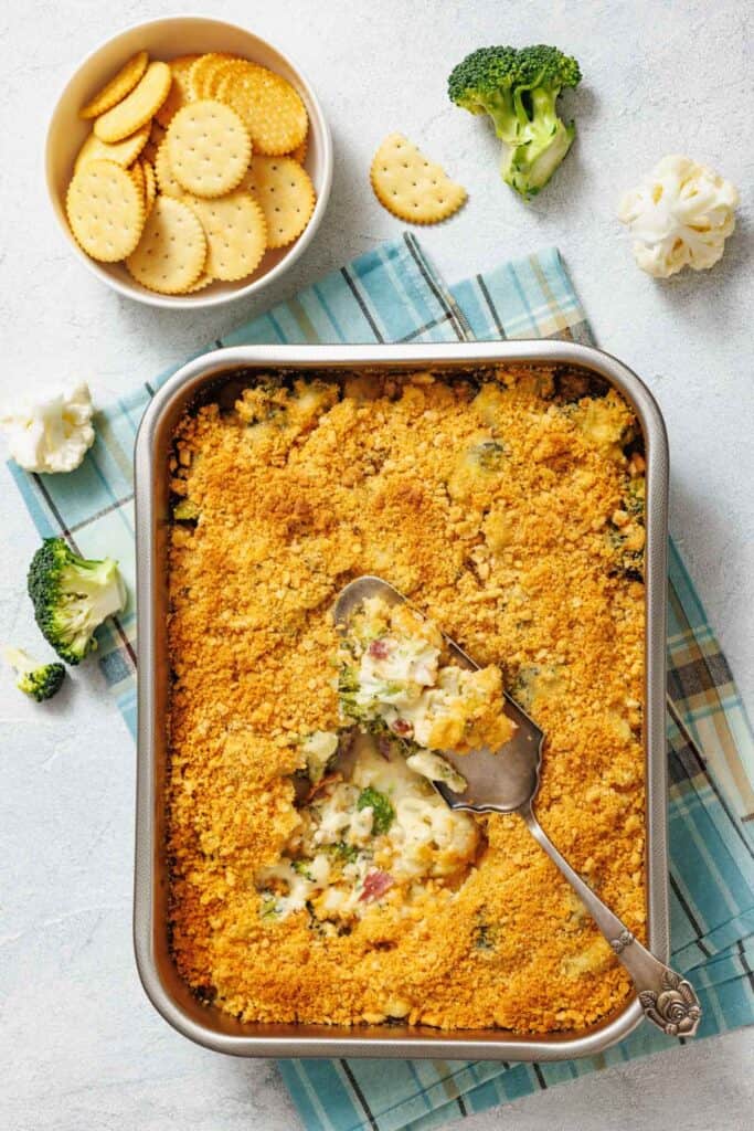 Cheesy vegetable casserole with a golden cracker topping, partially served in a baking dish.
