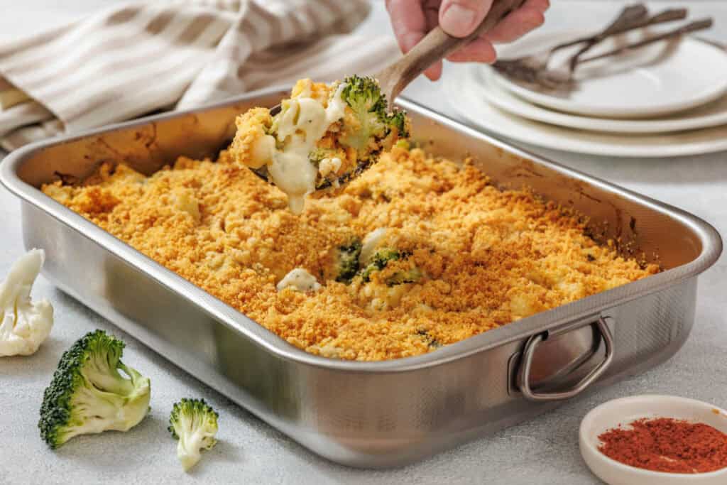A baked broccoli and cauliflower casserole with a golden breadcrumb topping, served in a metal baking dish with a wooden spoon lifting a creamy portion.