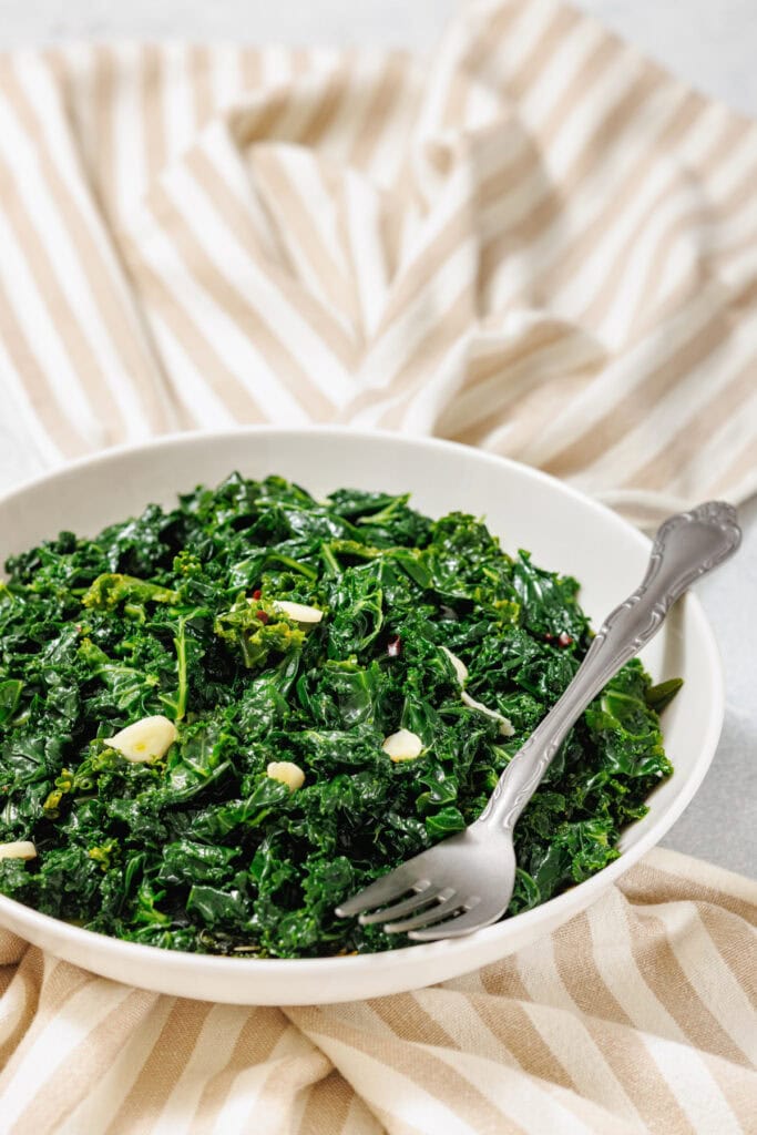 A white bowl filled with sautéed kale, glistening from light oil and garnished with thin slices of garlic. The leafy greens appear vibrant and tender, with a slightly wilted texture, served on a white plate over a light plaid tablecloth background.