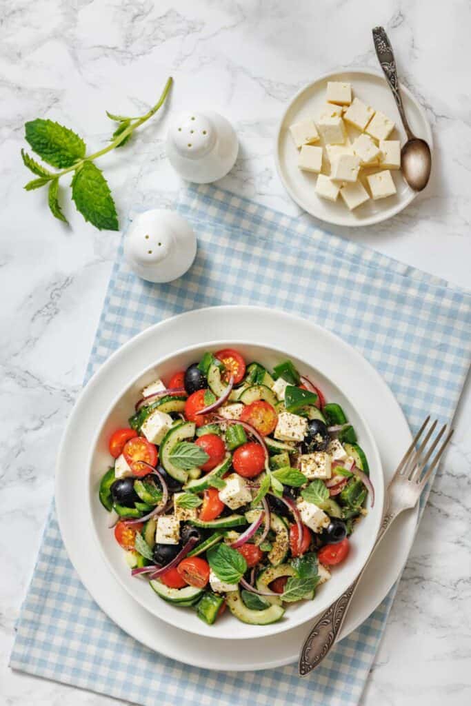 A Greek salad with cherry tomatoes, cucumber, olives, feta, and mint on a white plate, accompanied by extra feta and salt shakers on a blue gingham cloth.