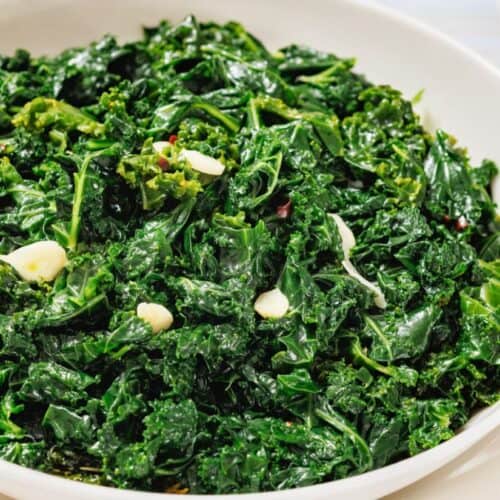 A white bowl filled with sautéed kale, glistening from light oil and garnished with thin slices of garlic. The leafy greens appear vibrant and tender, with a slightly wilted texture.