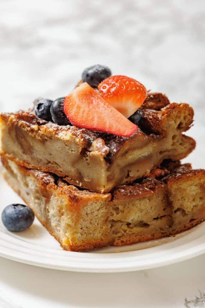 Close-up of two slices of bread pudding stacked on a white plate, topped with fresh strawberries and blueberries.