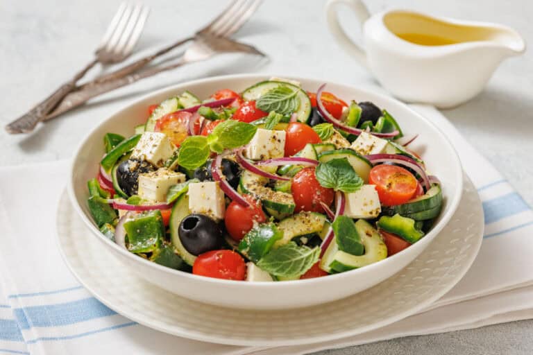 A fresh Greek salad served in a white bowl, featuring cherry tomatoes, cucumber slices, red onion, black olives, green bell peppers, and cubes of feta cheese, garnished with mint leaves and sprinkled with herbs.