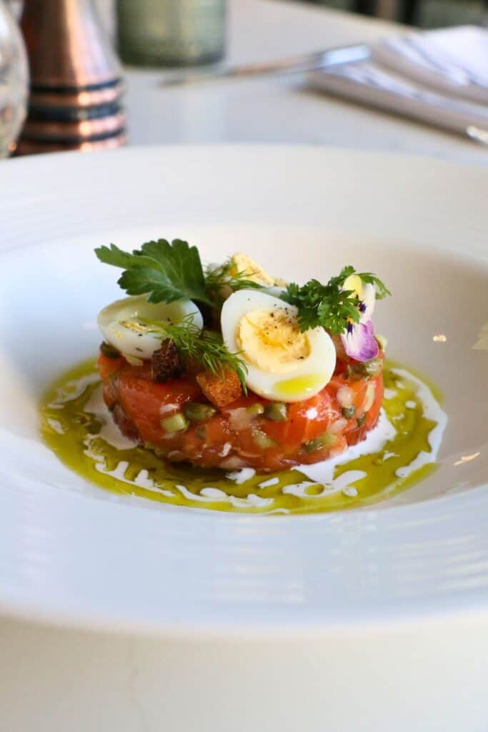 A refined dish of salmon tartare garnished with halved quail eggs, herbs, croutons, and edible flowers, surrounded by a swirl of chive oil and crème fraîche on a white plate.