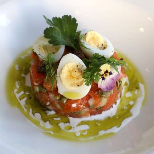 A gourmet dish of salmon tartare topped with halved quail eggs, herbs, and edible flowers, surrounded by a drizzle of chive oil and crème fraîche on a white plate.