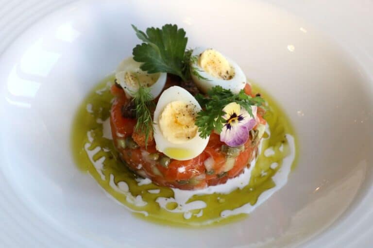 A gourmet dish of salmon tartare topped with halved quail eggs, herbs, and edible flowers, surrounded by a drizzle of chive oil and crème fraîche on a white plate.