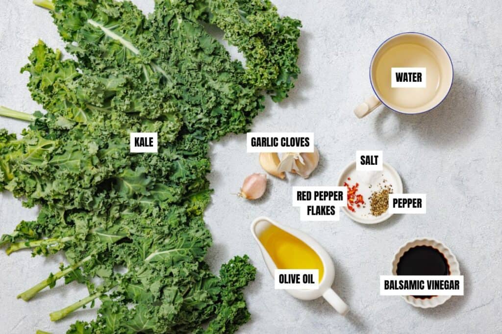 A flat lay of ingredients for sautéed kale, labeled clearly on a light surface. Fresh curly kale is spread out on the left. To the right are garlic cloves, a cup of water, a dish with red pepper flakes, salt, and black pepper, a small pitcher of olive oil, and a small bowl of balsamic vinegar.