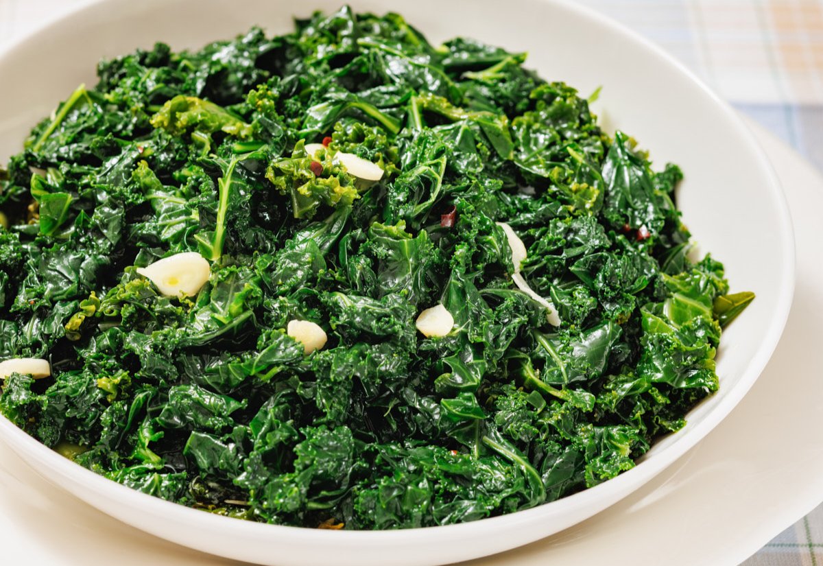 A white bowl filled with sautéed kale, glistening from light oil and garnished with thin slices of garlic. The leafy greens appear vibrant and tender, with a slightly wilted texture.