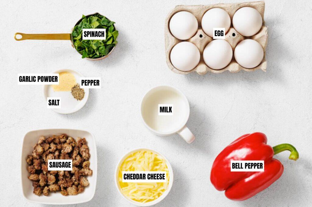 Ingredients for a recipe are neatly arranged on a white surface, including spinach, eggs, garlic powder, pepper, salt, milk, sausage, cheddar cheese, and a red bell pepper.