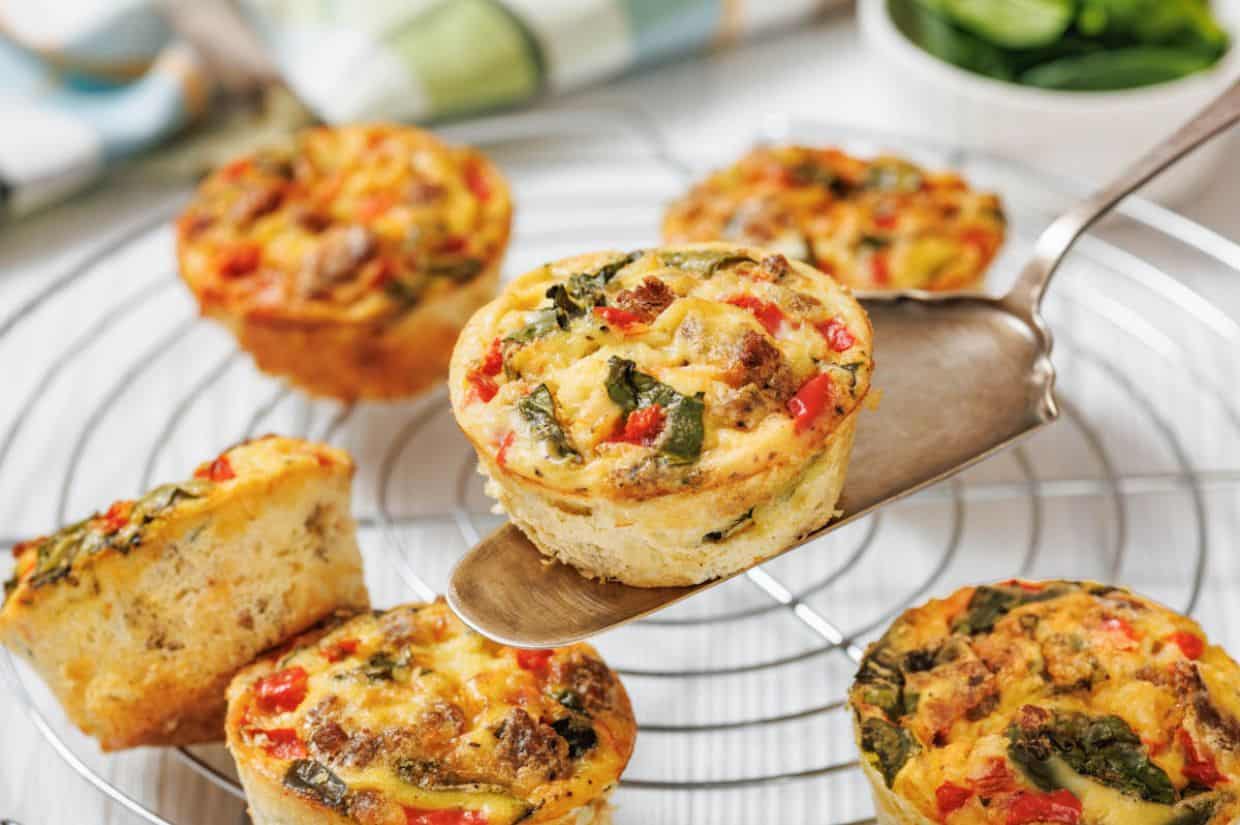 Close-up of savory muffins topped with spinach, red peppers, and sausage pieces on a wire rack. One muffin is lifted with a spatula, conveying a fresh and appetizing feel.