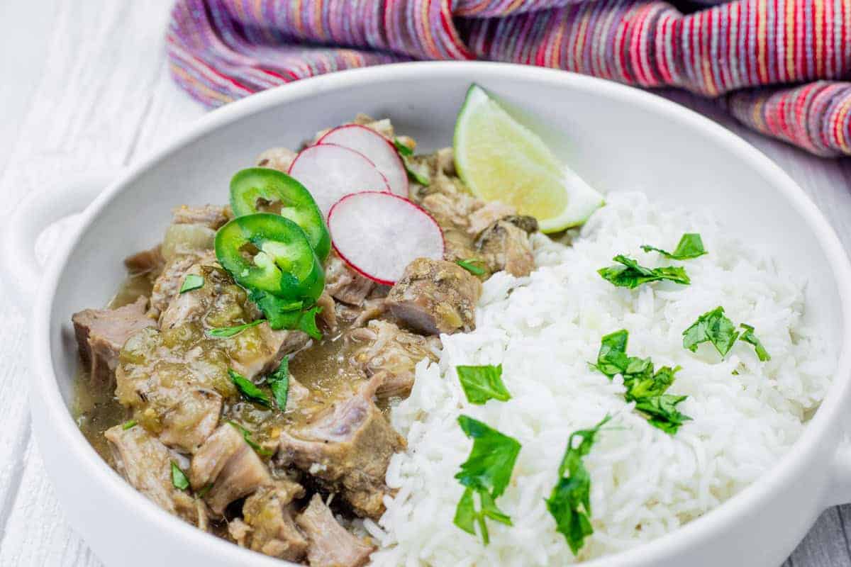 A bowl of white rice and green chili verde garnished with cilantro, radish slices, jalapeño, and a lime wedge, set on a white surface with a colorful cloth in the background.
