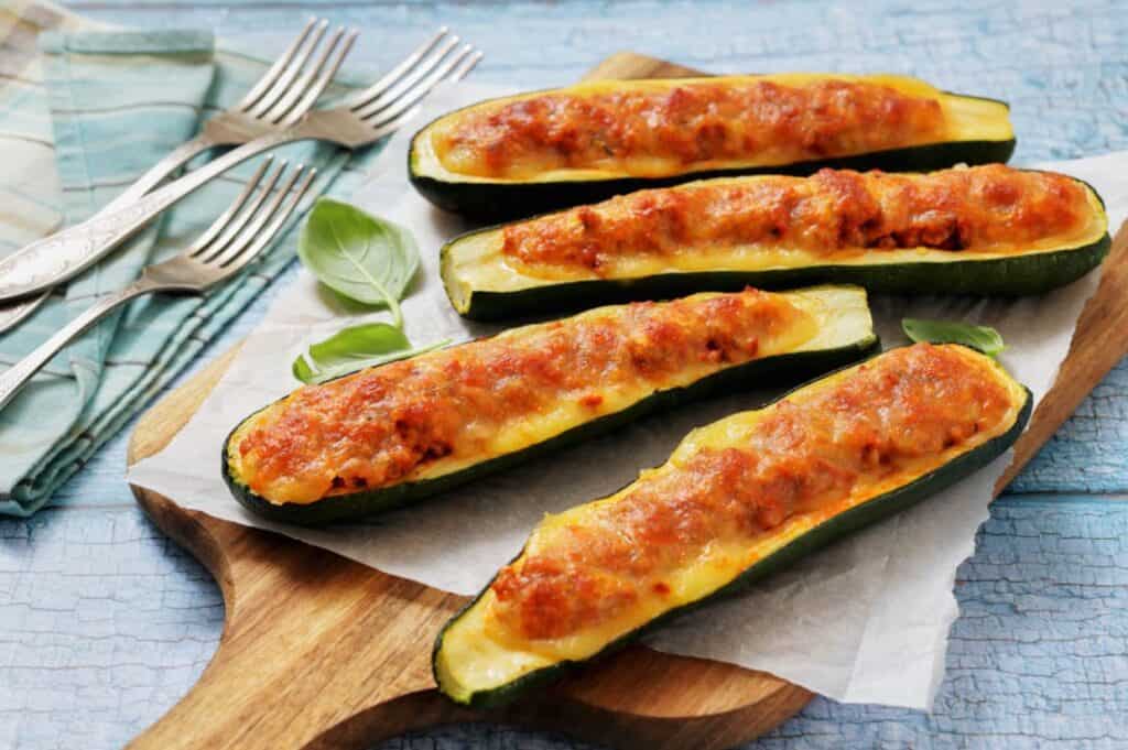 Baked zucchini boats stuffed with a rich meat and cheese topping on a wooden board. Four silver forks rest on a blue patterned cloth nearby.