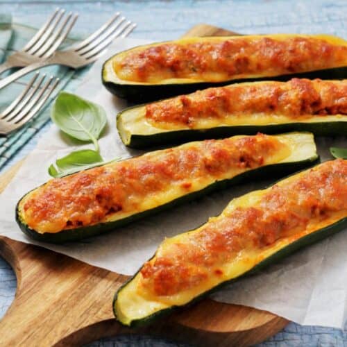 Baked zucchini boats stuffed with a rich meat and cheese topping on a wooden board. Four silver forks rest on a blue patterned cloth nearby.