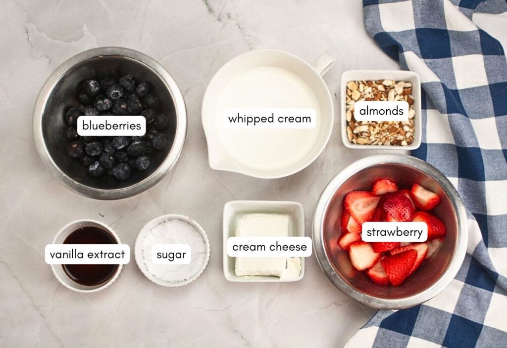 A marble surface displays ingredients for a dessert: bowls of blueberries, sliced strawberries, chopped almonds, cream cheese, sugar, vanilla extract, and a measuring cup of whipped cream, each labeled with white tags.