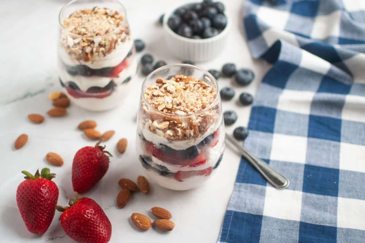 Two parfait glasses filled with layered yogurt, strawberries, blueberries, and nuts. Nearby are fresh almonds, strawberries, blueberries, and a blue checkered cloth.