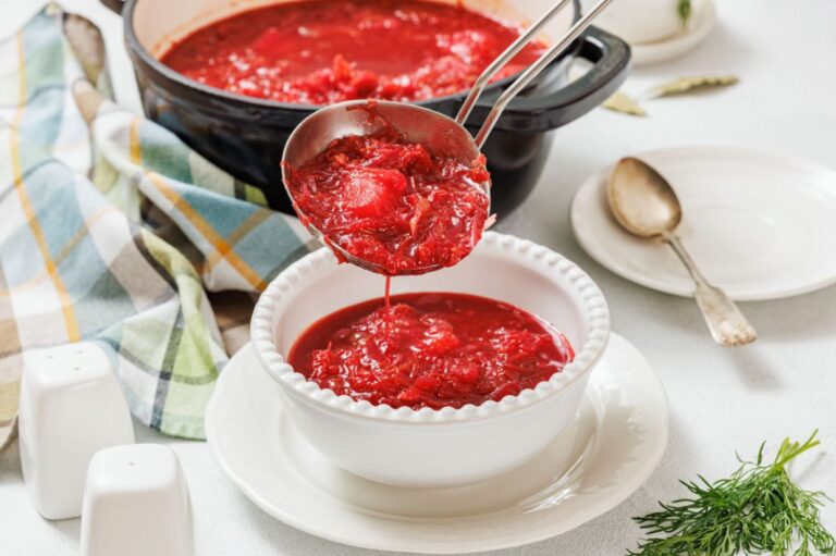A ladle pours vibrant red borscht into a white bowl, set on a table with a striped cloth and dill garnish. A rustic, cozy kitchen vibe is conveyed.