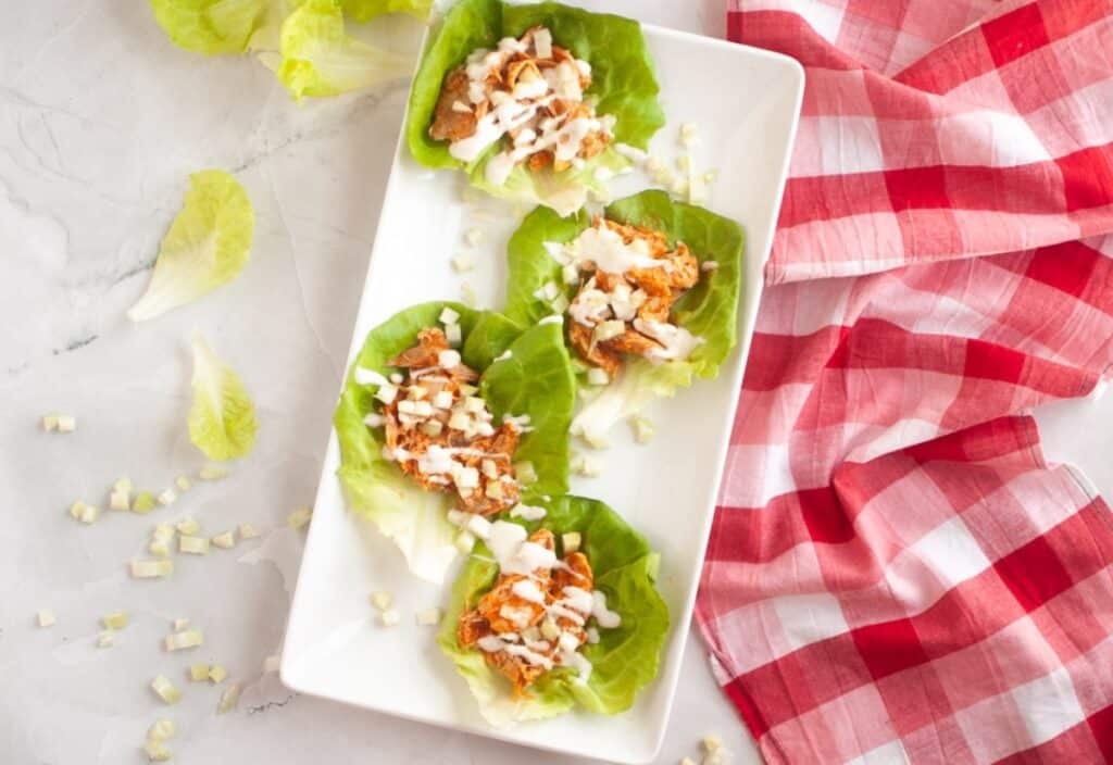 Four lettuce wraps filled with shredded chicken and dressing on a white plate, surrounded by fresh lettuce and a red checkered cloth. Cream dressing in a small bowl nearby.