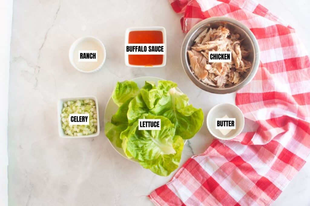 Ingredients for a buffalo chicken lettuce wrap on a marble surface: ranch dressing, buffalo sauce, shredded chicken, celery, lettuce, butter.
