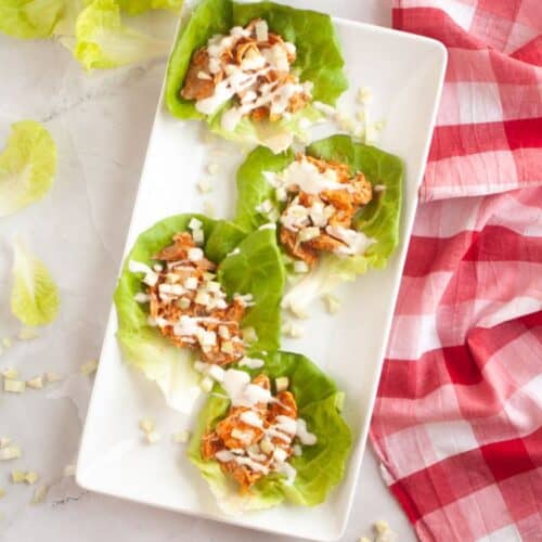 Four lettuce wraps with shredded meat and diced onions, drizzled with white sauce, are neatly arranged on a white rectangular plate. A red checkered cloth decorates the side, conveying a fresh and inviting tone.