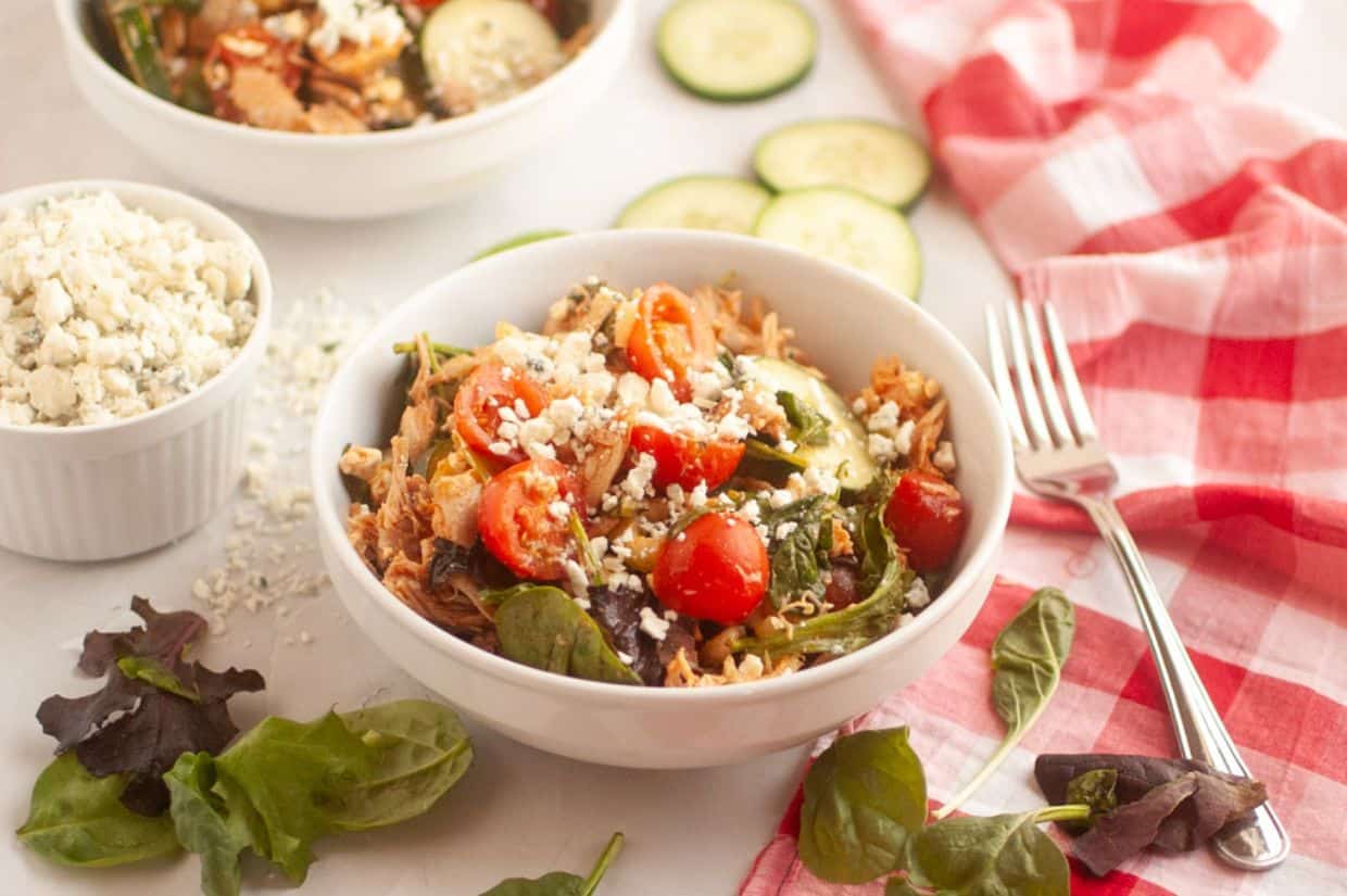 Bowl of salad with cherry tomatoes, cucumbers, leafy greens, and crumbled cheese on a white surface. A red checkered napkin and fork nearby.