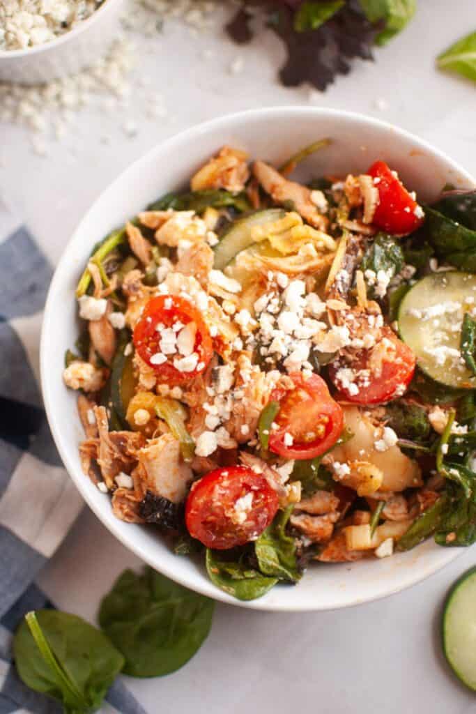 A colorful salad in a white bowl featuring cherry tomatoes, spinach, cucumber slices, shredded chicken, and crumbled feta cheese, conveying freshness.