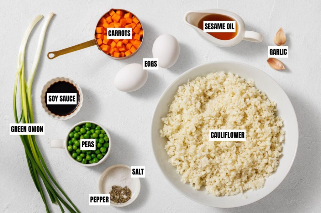 Ingredients for cauliflower fried rice on a white surface: a bowl of riced cauliflower, chopped carrots, eggs, sesame oil, garlic, green onion, soy sauce, peas, salt, and pepper.