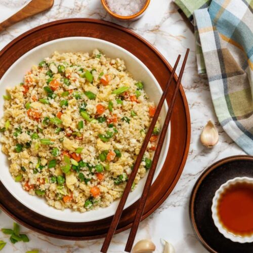 A bowl of cauliflower fried rice with chopped vegetables and scrambled eggs, garnished with green onions.