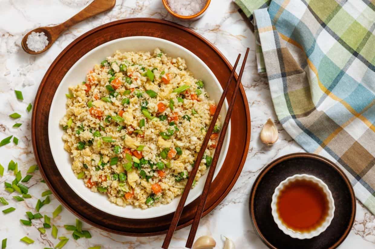 Ingredients for cauliflower fried rice on a white surface: a bowl of riced cauliflower, chopped carrots, eggs, sesame oil, garlic, green onion, soy sauce, peas, salt, and pepper.