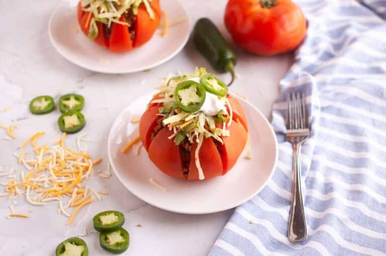 Two taco-stuffed tomatoes on white plates with toppings and a fork.