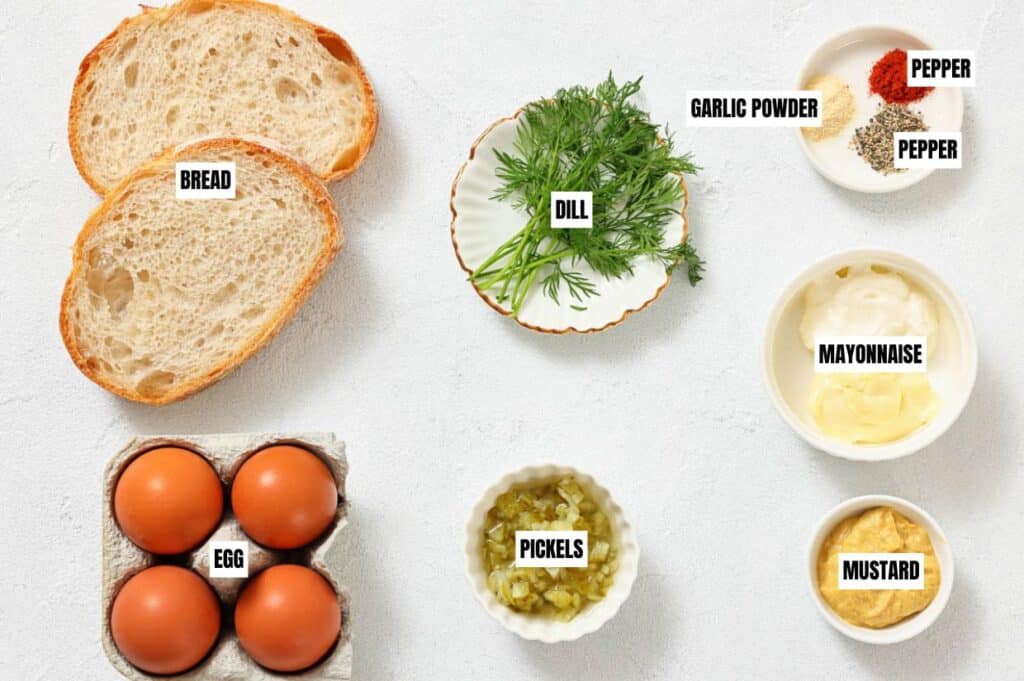 Two slices of bread, fresh dill, garlic powder, black pepper, mayonnaise, mustard, pickles, and four brown eggs on a light background. Ingredients labeled.