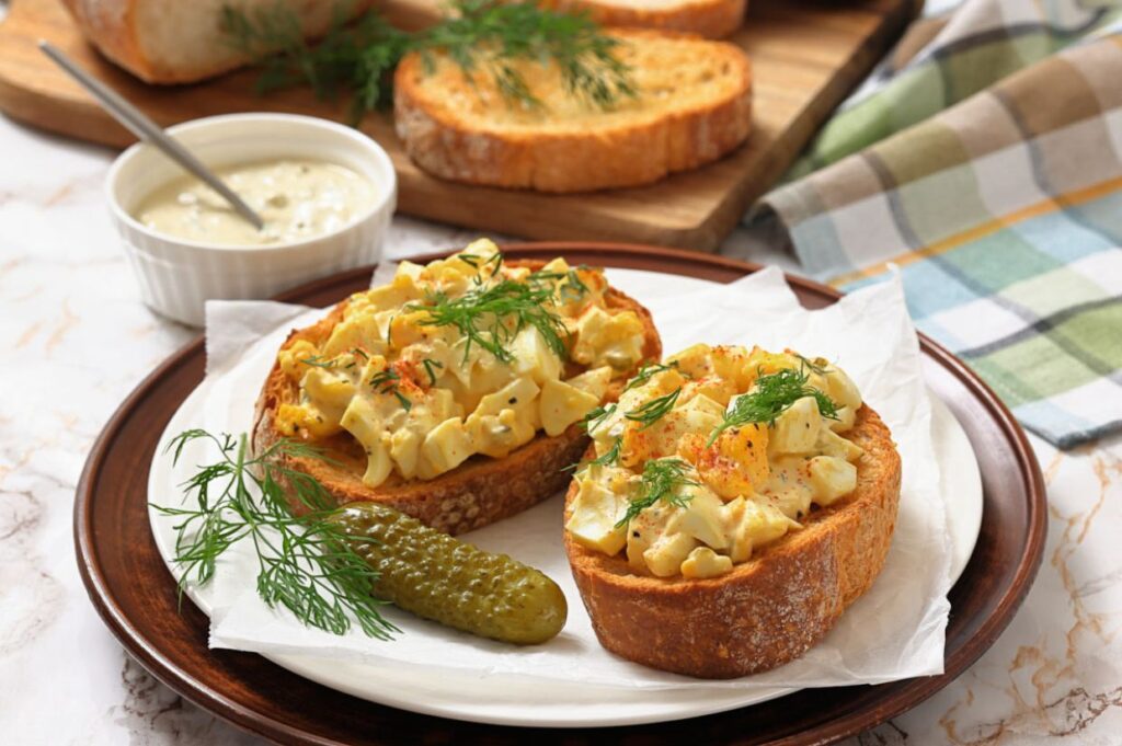 Two slices of toasted bread topped with creamy deviled egg salad and fresh dill, served with a pickle on the side.