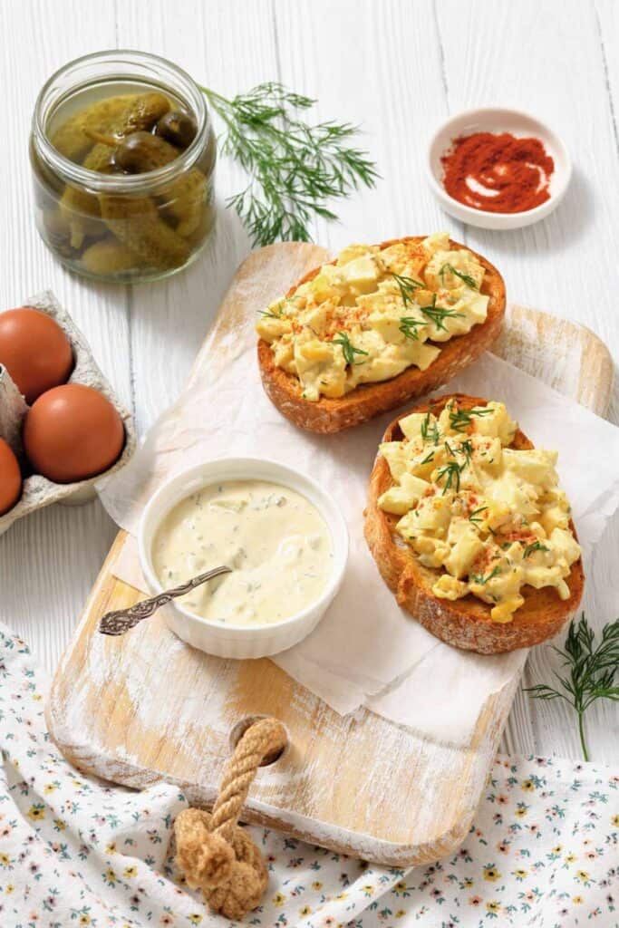 Two slices of toast topped with creamy egg salad, garnished with dill and paprika, on a wooden board with a bowl of dressing, pickles, and eggs nearby.