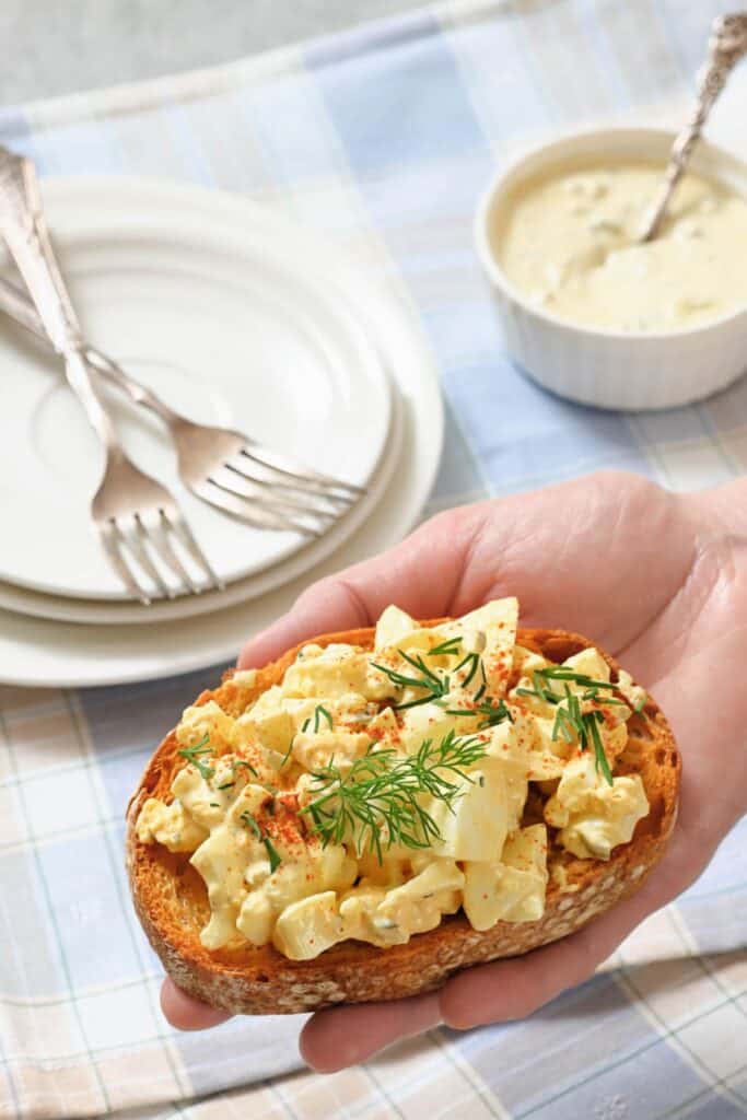 Hand holding egg salad on toasted bread, garnished with dill and paprika. A nearby table holds stacked plates and forks, next to a small dish. Cozy and appetizing.