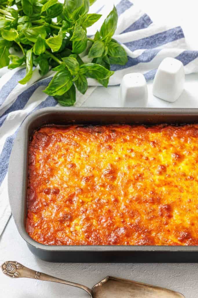 Golden-brown baked casserole in a dark pan on a striped towel, garnished with fresh basil leaves. A silver serving spoon lies beside it.
