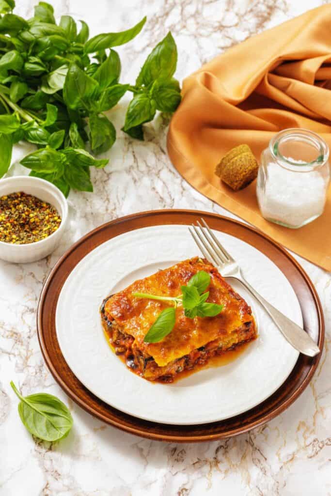 A slice of eggplant parmigiana topped with fresh basil on a white plate with a brown rim. Surrounded by basil leaves, a spice bowl, salt jar, and orange cloth.