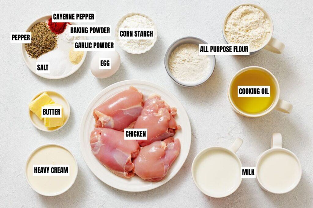 A neatly arranged set of ingredients for a recipe. Chicken pieces center, surrounded by butter, spices, egg, flours, cream, milk, and oil on a light surface.