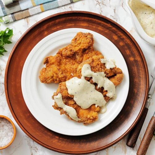 Crispy fried chicken smothered in creamy white gravy on a white plate with a brown rim. Side of fresh lettuce, salt, cutlery, and sauce on marble surface.