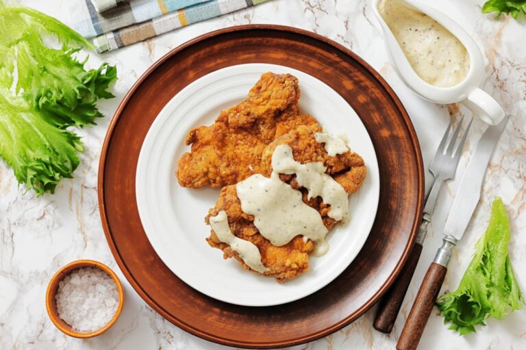 Crispy fried chicken smothered in creamy white gravy on a white plate with a brown rim. Side of fresh lettuce, salt, cutlery, and sauce on marble surface.