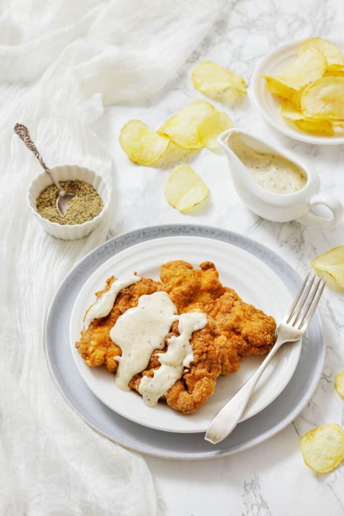 A crispy, golden chicken fried steak with creamy white gravy on a white plate. Surrounded by potato chips, gravy boat, spices, and a fork.